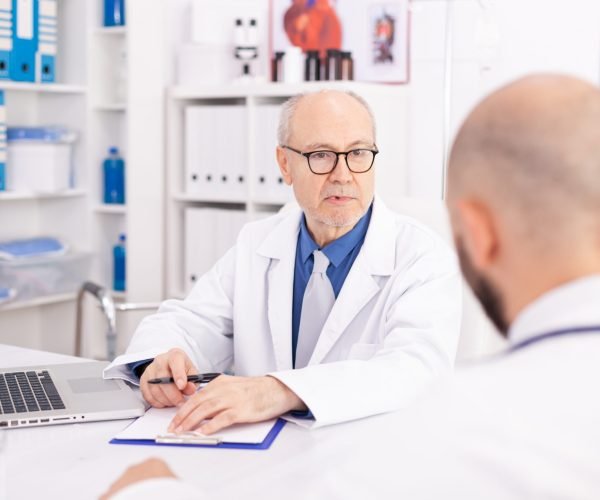 Experienced mature doctor explaining patient diagnosis to young medical physician during conference. Clinic expert therapist talking with colleagues about disease, medicine professional.