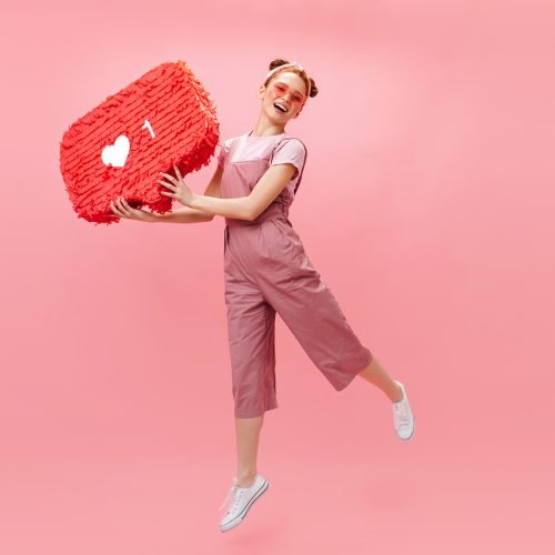 Joyful woman in stylish jumpsuit jumping on pink background, holding like sign