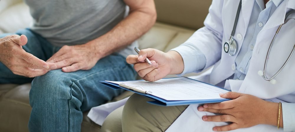 Crop shot of senior man sitting on sofa with doctor during home visit taking notes on clipboard
