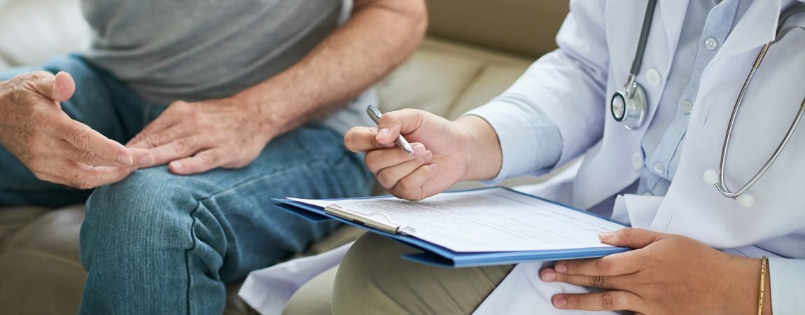 Crop shot of senior man sitting on sofa with doctor during home visit taking notes on clipboard